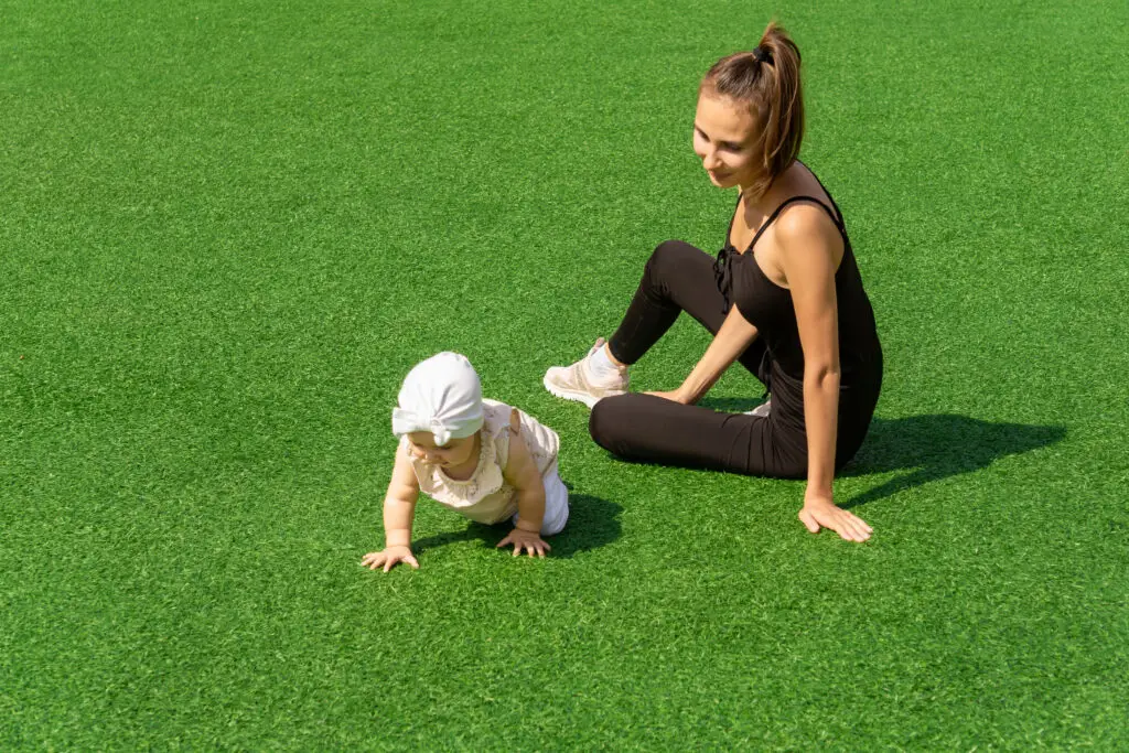 Mother sitting on clean artificial turf while her young child crawls safely across the grass in a backyard.